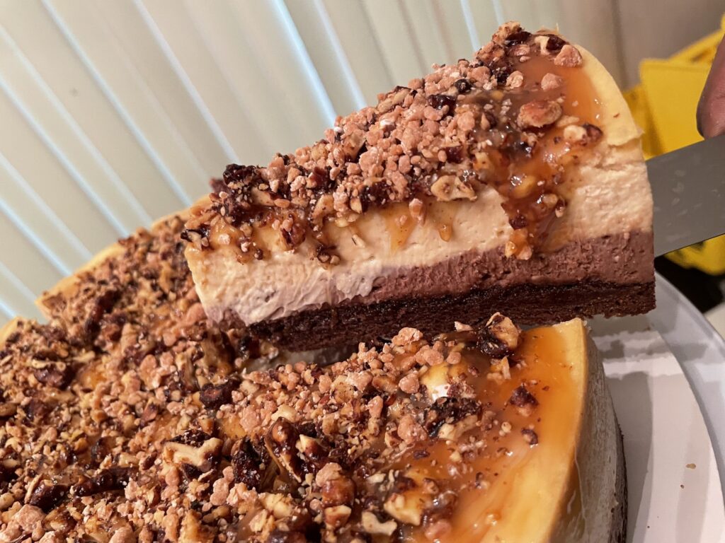 A lifted slice of cheesecake with a chocolate base, caramel-colored topping and chopped nuts above the remaining cake.