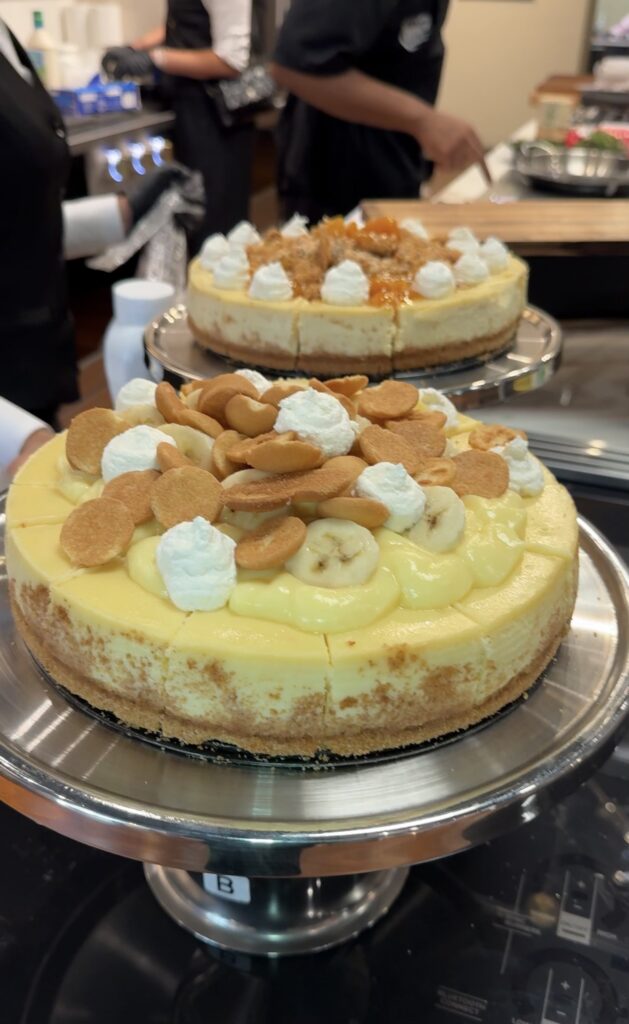 Two cheesecakes on silver stands, topped with banana slices, vanilla wafer cookies and piped cream.